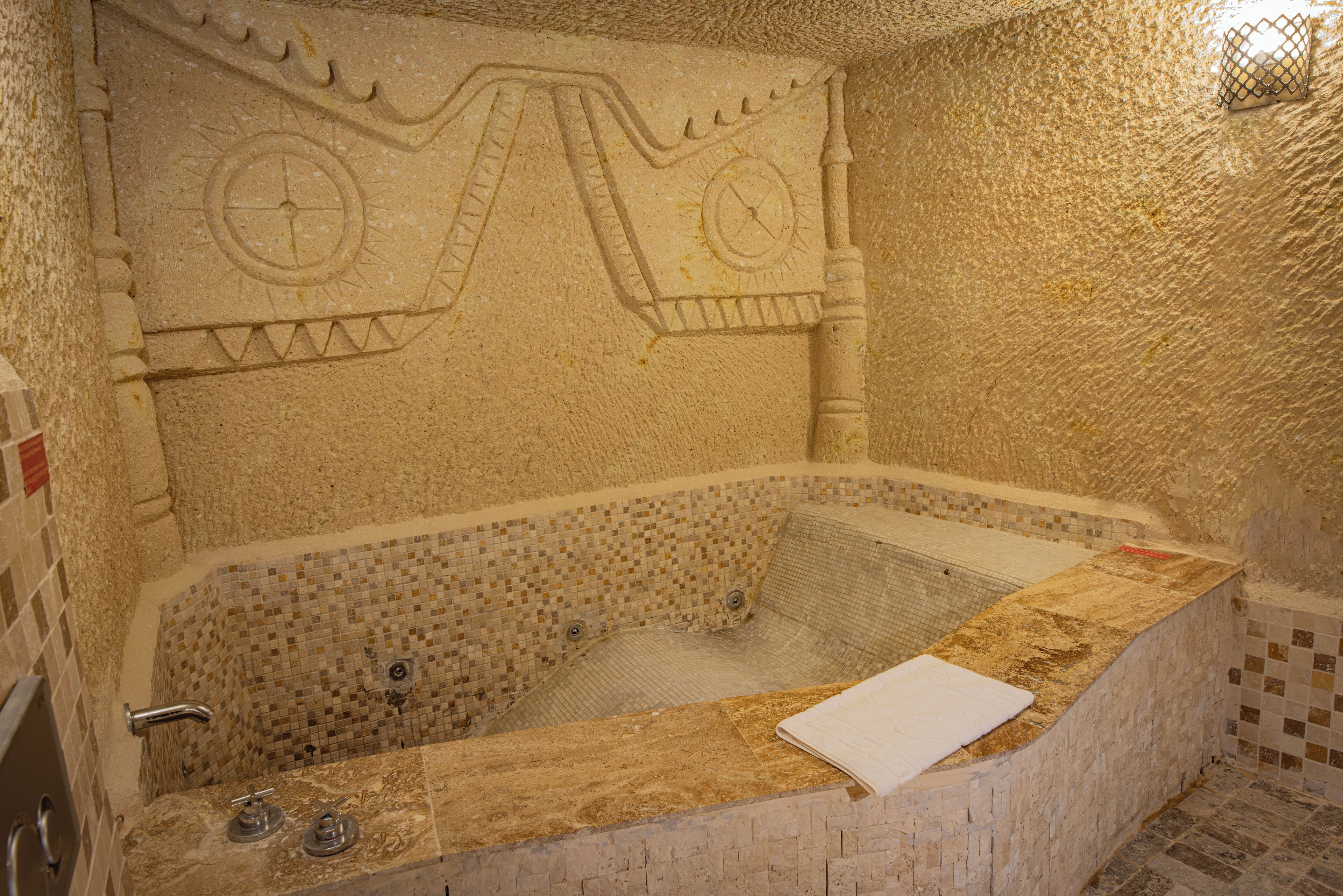 A stone bathtub with mosaic tile walls and a white towel on the edge.