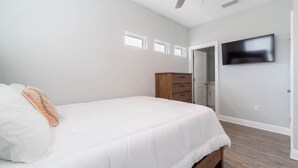 Una camera da letto con un letto, una cassettiera in legno, una ventola a soffitto, una TV fissata al muro e una porta che dà su un'altra stanza.