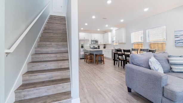 Un escalier mène à une cuisine dotée de meubles blancs, à un coin repas avec un canapé gris et offre une vue sur l’extérieur grâce à de grandes fenêtres.