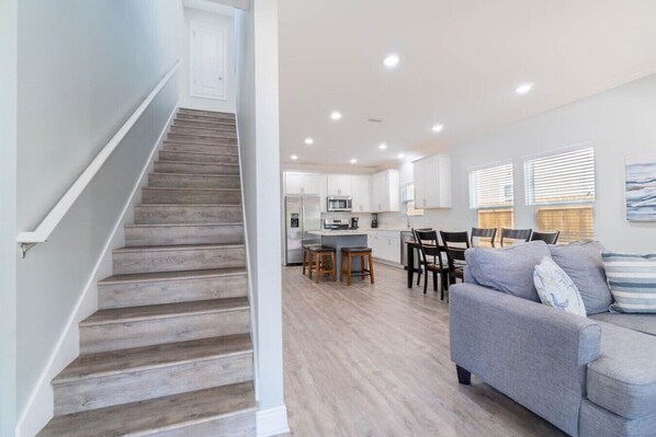 Un escalier mène à une cuisine dotée de meubles blancs, à un coin repas avec un canapé gris et offre une vue sur l’extérieur grâce à de grandes fenêtres.