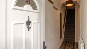 Une porte blanche avec un panneau en verre et un heurtoir en métal, un escalier recouvert d’une moquette à motifs et un luminaire suspendu.