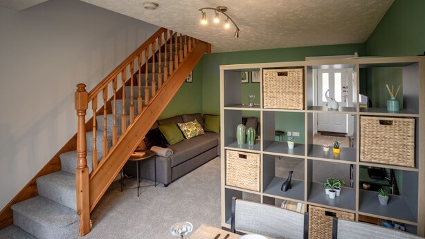 Een woonkamer met een trap, een grijze bank, een eettafel gedekt met borden en glazen, en een inbouwkast met manden.