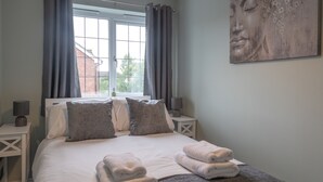 Een net opgemaakt bed met kussens en opgevouwen handdoeken, een raam met gordijnen, een bedlamp en een wanddecoratie.