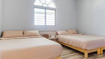 A bedroom with two beds, a window with blinds, a small table with a basket and tissues, and a wooden floor.
