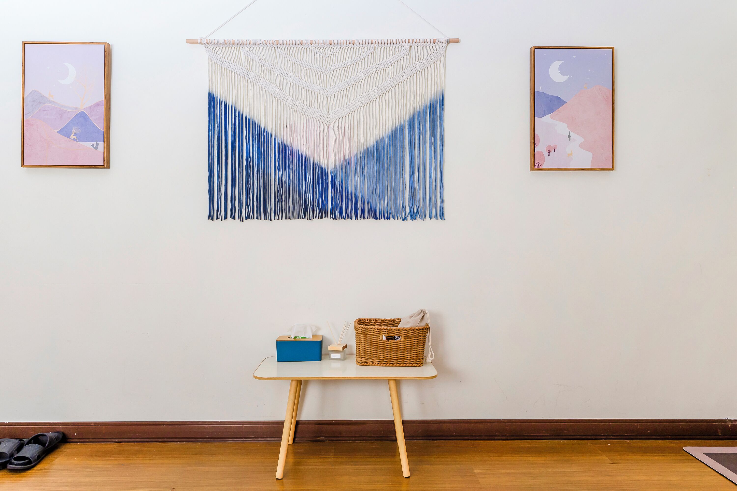 A room with a wooden floor, a small table with a basket and a blue box, and two framed artworks on the wall.