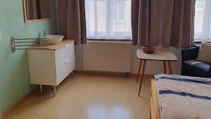 Quarto com cama, uma mesinha, radiador, janela com cortinas, espelho e pia.
