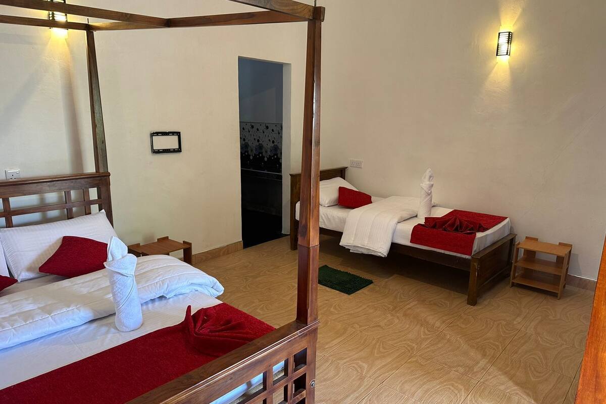A four-poster bed with white linens and red accents, two single beds, wooden flooring, and a small wooden side table.