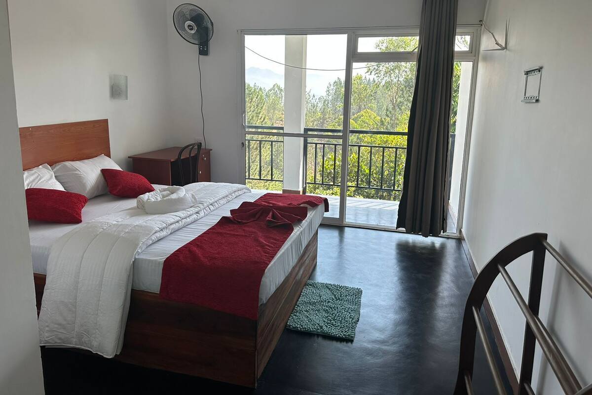 A bedroom with a bed, a nightstand, a fan, and a view of greenery through a large window.