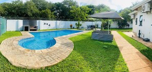 A backyard with a swimming pool, a covered patio area, and a white building. - Murugare Lodge (Victoria Falls)