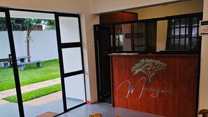 Un'area di accoglienza con una reception in legno che presenta un logo raffigurante un albero e il nome 'Myrigare'. Si apre una porta in vetro verso l'esterno, ed è presente una panca in legno con quotidiani sopra.