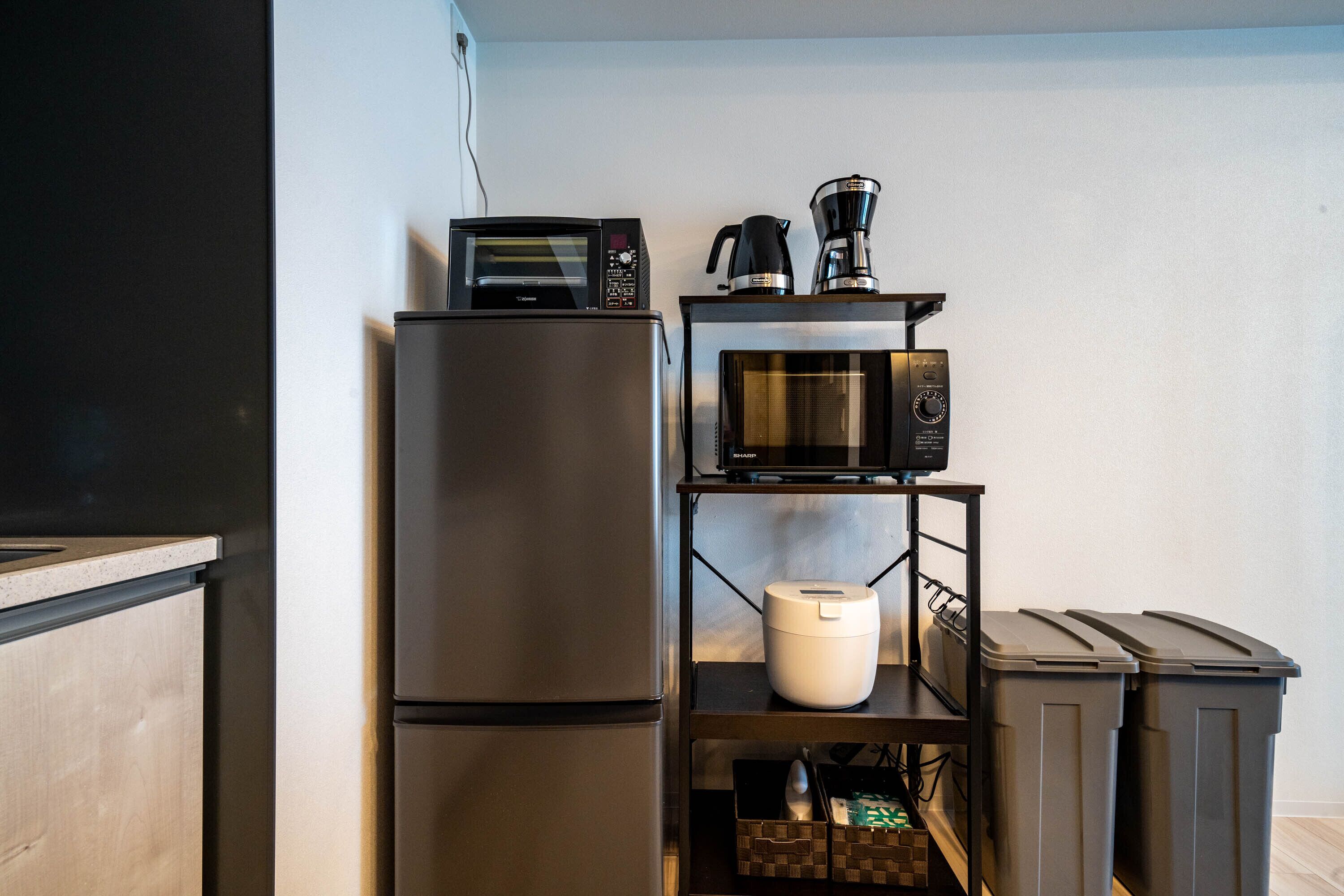 Cocina compacta con refrigerador, microondas, cafetera y un estante pequeño con un recipiente blanco y botes de basura.