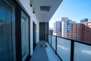 A narrow balcony with a glass door, a small mat, and a view of tall buildings in the background. - MOXIE 603 (Sapporo)