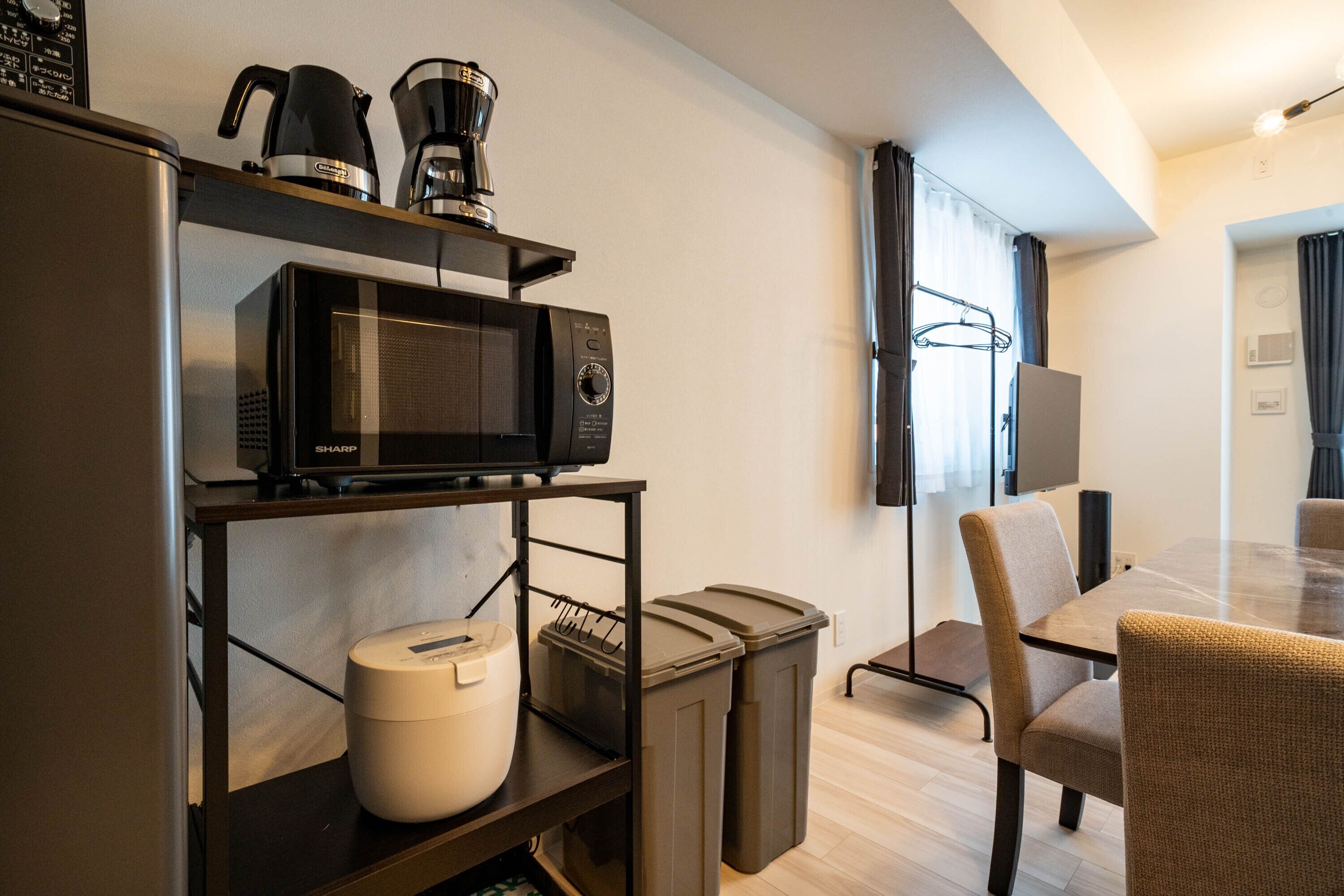 Habitación de hotel moderna con microondas, cafetera y un electrodoméstico blanco sobre una estantería. Incluye una mesa de comedor con sillas y un televisor montado en la pared.