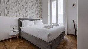 A modern bedroom with a grey bed, white bedding, a herringbone wooden floor, a white nightstand, and a wall with a geometric pattern.