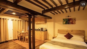A bedroom with a wooden bed, a nightstand, and a desk with a chair. There is a kitchen area with cabinets and a microwave. The room has wooden beams on the ceiling and a painting on the wall. - Wittys hotel (Lalitpur)