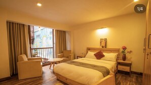 A hotel room with a large bed, a chair, a small table, and a view of a colorful building through the window. - Wittys hotel (Lalitpur)