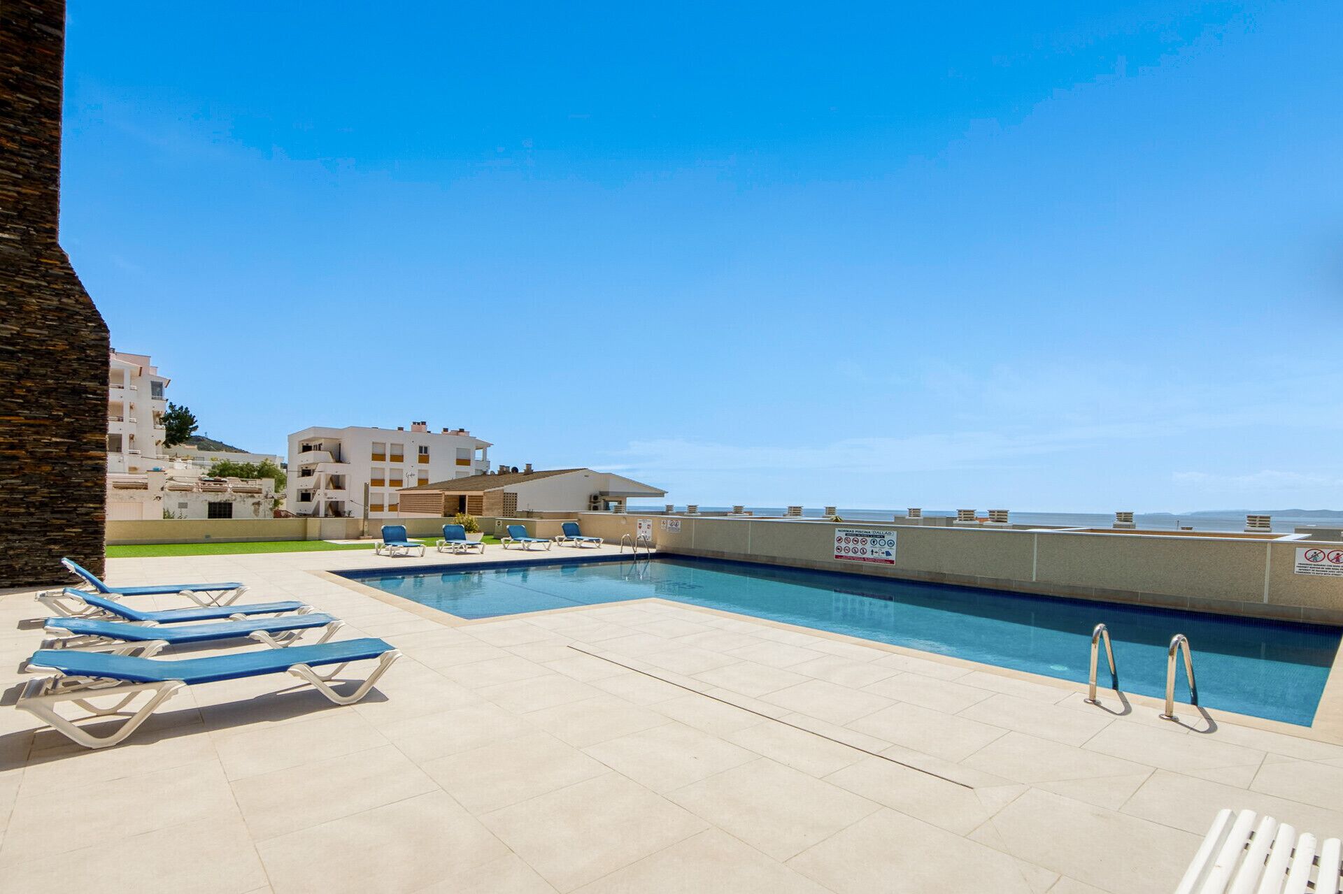 A swimming pool area with lounge chairs, a clear sky, and buildings in the background.