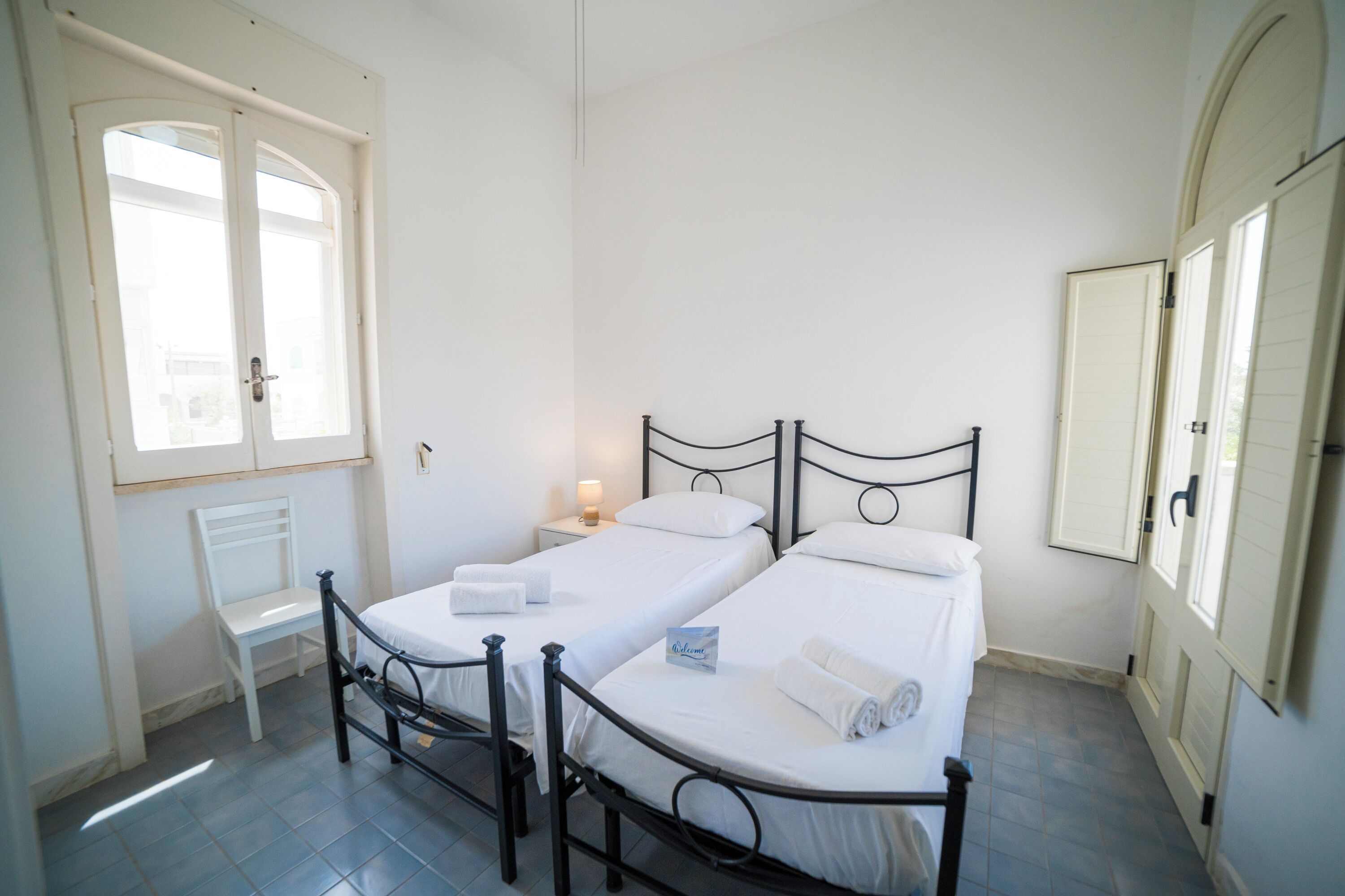 Une chambre avec deux lits simples, une chaise, une table de chevet avec une lampe et une fenêtre.