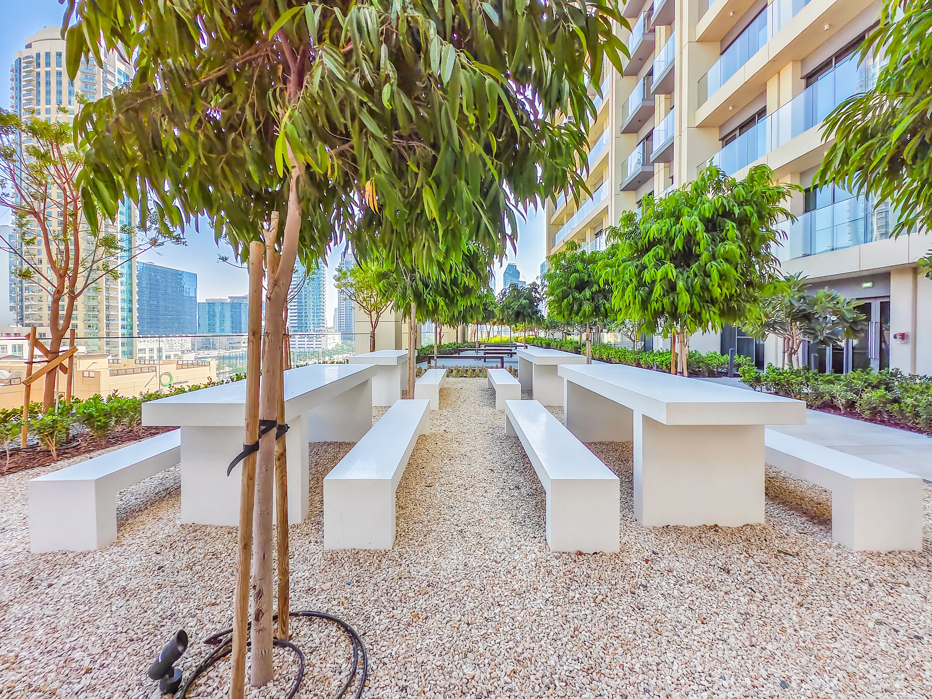 Un espace extérieur moderne aménagé avec des bancs blancs et des arbres, entouré d’un chemin de gravier et de bâtiments en arrière-plan.