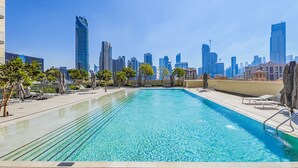 A rooftop pool with clear blue water, surrounded by lounge chairs and umbrellas, set against a backdrop of modern high-rise buildings. - Cityscape Splendor Lavish 1BR Uptown  (Dubai)