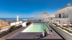 Una piscina sul tetto con vista chiara sul mare, un edificio bianco e un pergolato con posti a sedere.