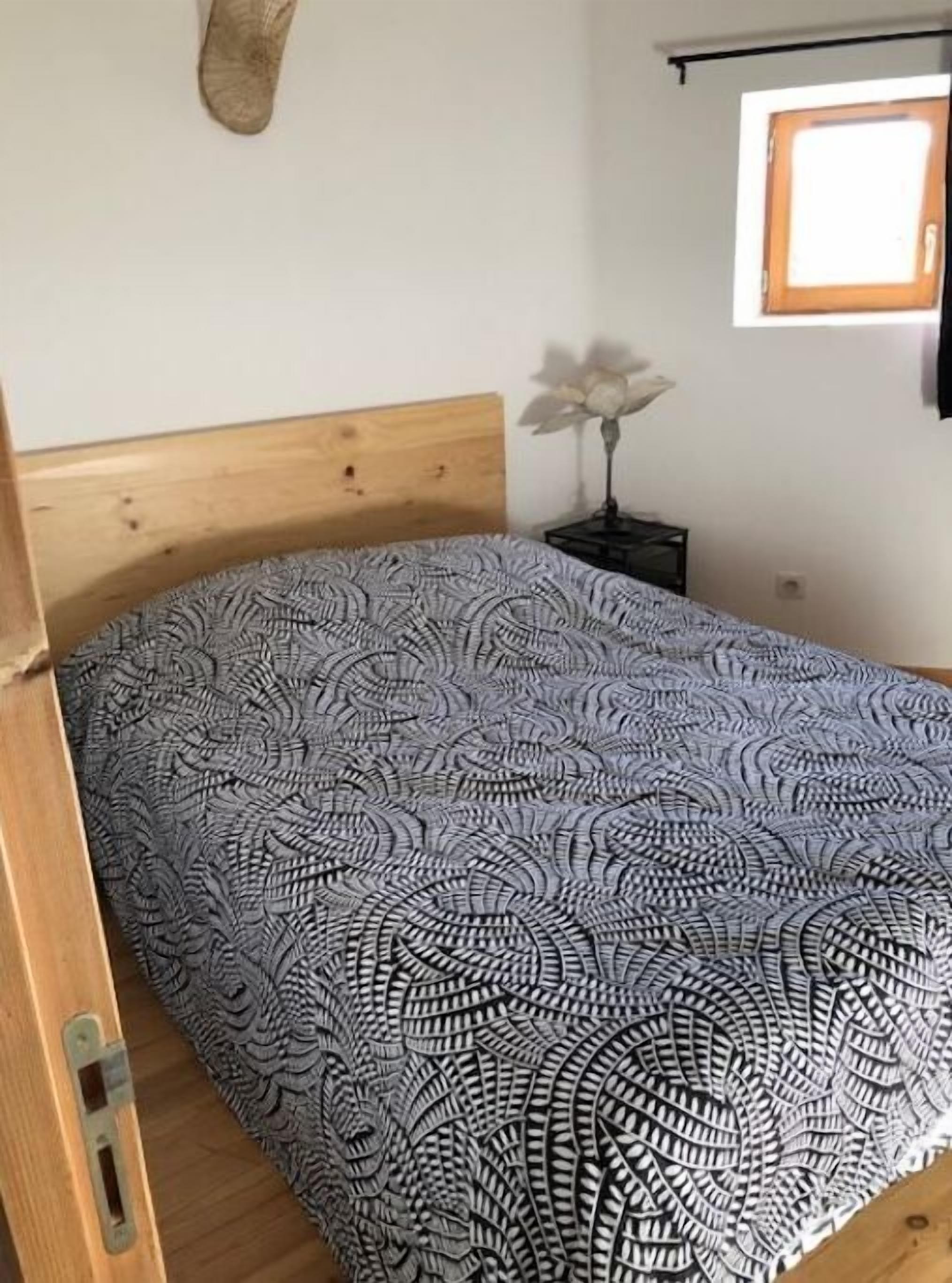 Een bed met een geborduurde spreideken, een houten hoofdbord, een klein nachtkastje met een lamp en een raam met houten kader.