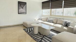 A modern living room with a sectional sofa, a coffee table, and a large window with blinds. - Black White Modern Spacious (Tunis)