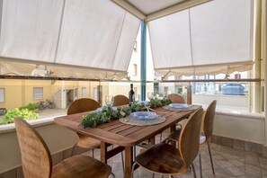 An outdoor dining area with a wooden table set for four, surrounded by chairs, under a large canopy. - Sardinia Re - Malta 29 Apartment (Alghero)