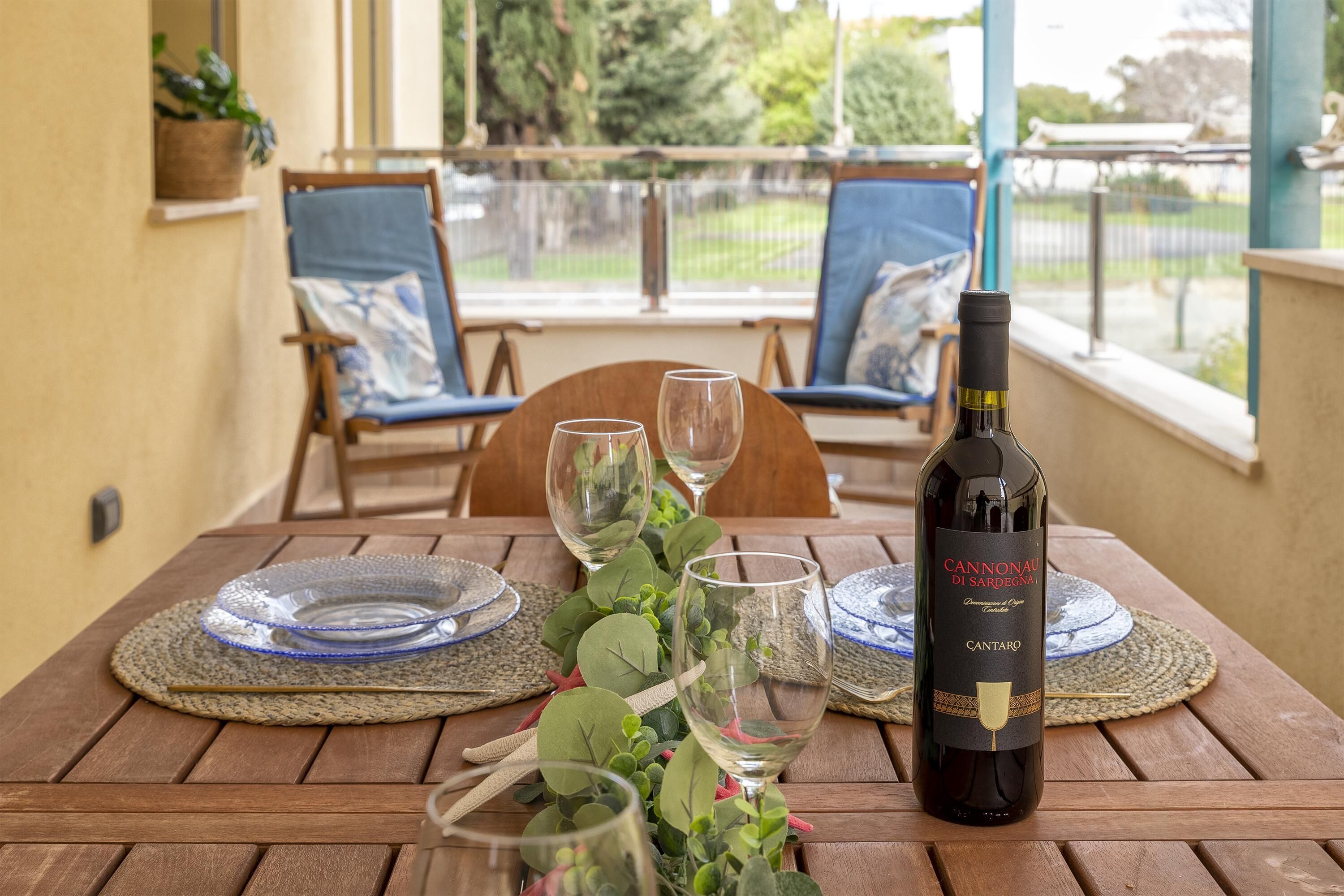 Una mesa de madera preparada para una comida, con una botella de vino, dos sillas y vista a un jardín desde una ventana.