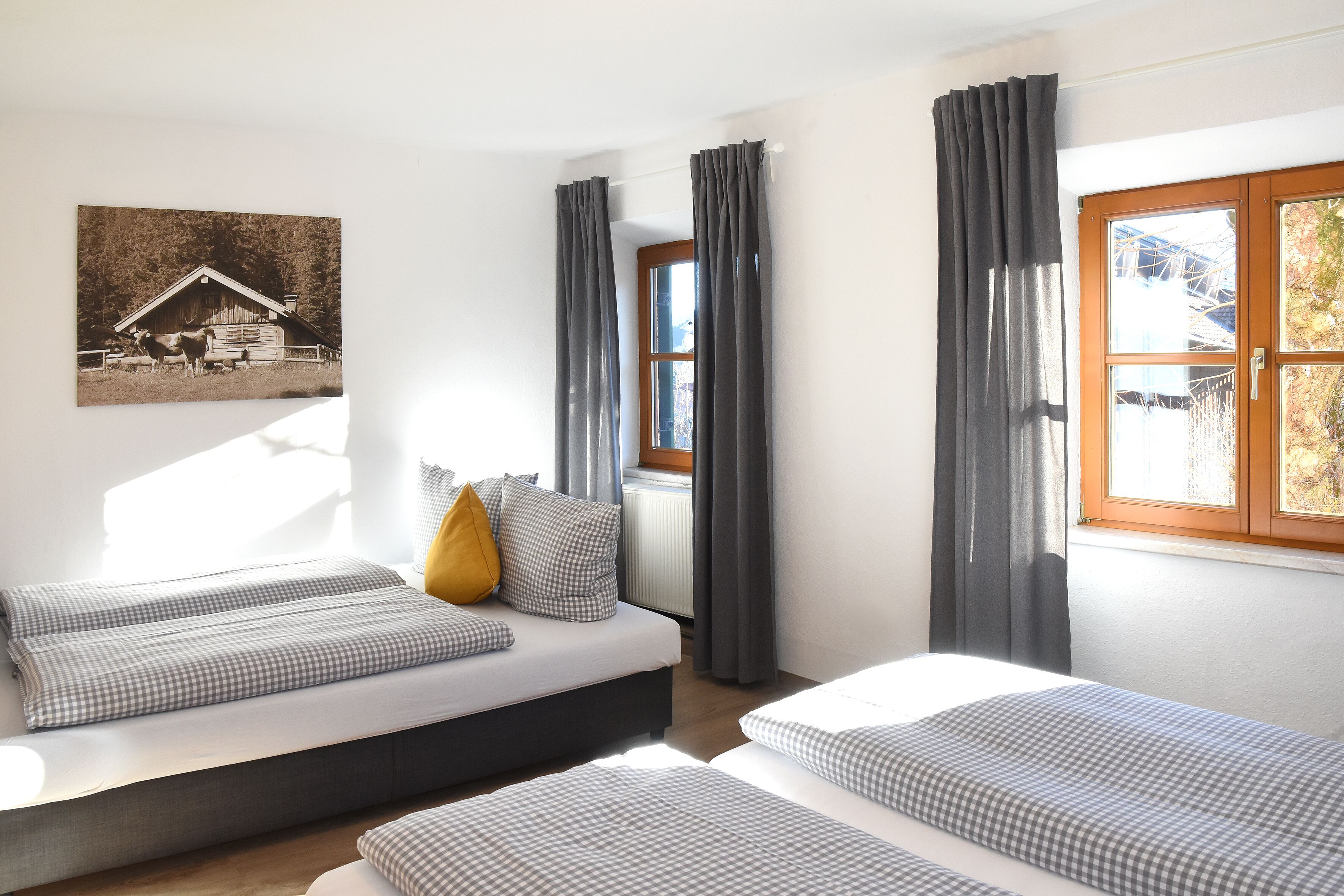Una habitación con dos camas, una ventana con cortinas, un radiador y un cuadro colgado en la pared que representa un granero.