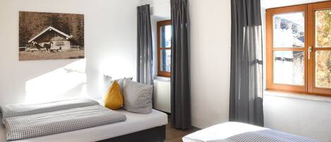 Una habitación con dos camas, una ventana con cortinas, un radiador y un cuadro colgado en la pared que representa un granero.