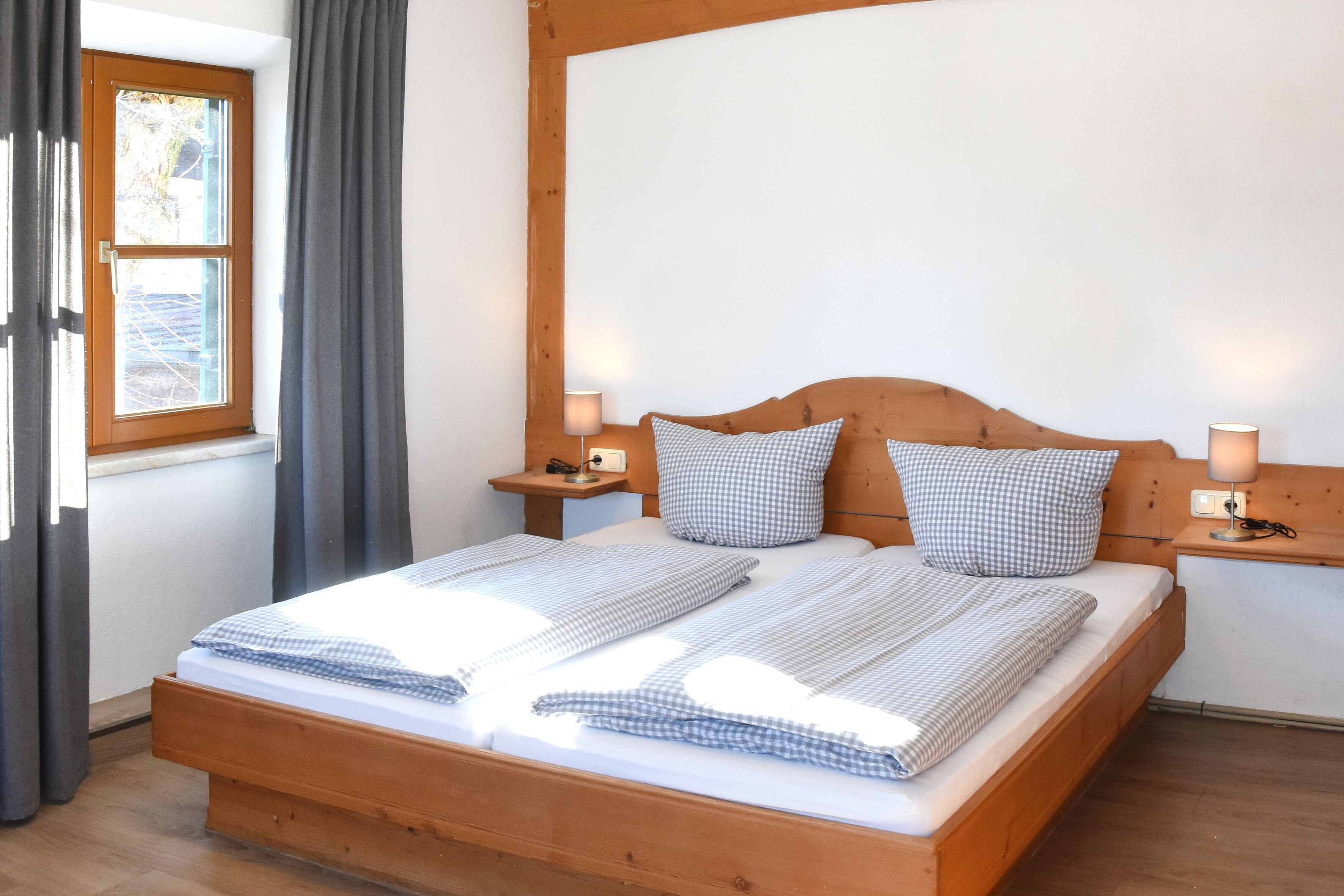 A neatly made bed with checkered bedding, wooden headboard, bedside lamps, and a window with curtains.