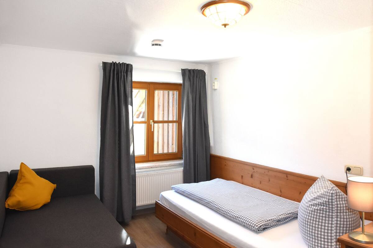 A bedroom with a bed, a sofa, a window with curtains, a lamp, and a radiator.