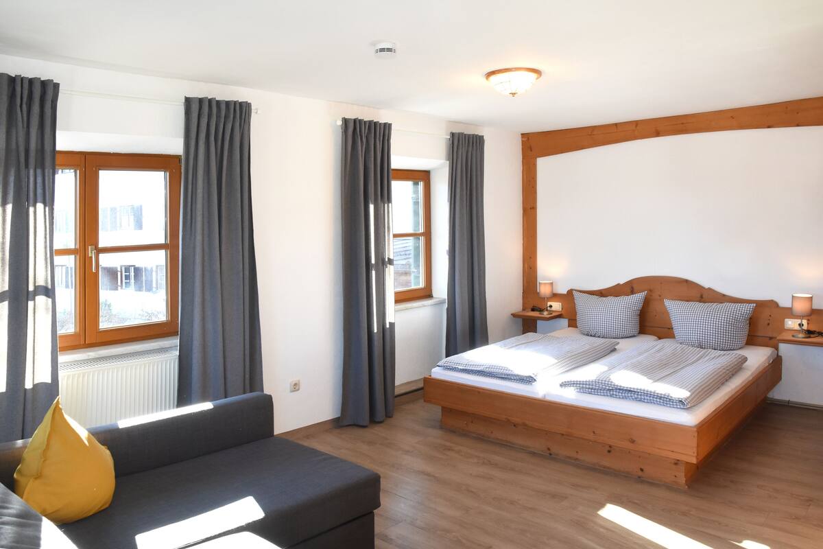 A bedroom with a wooden bed, grey curtains, a sofa, and a window with a view.