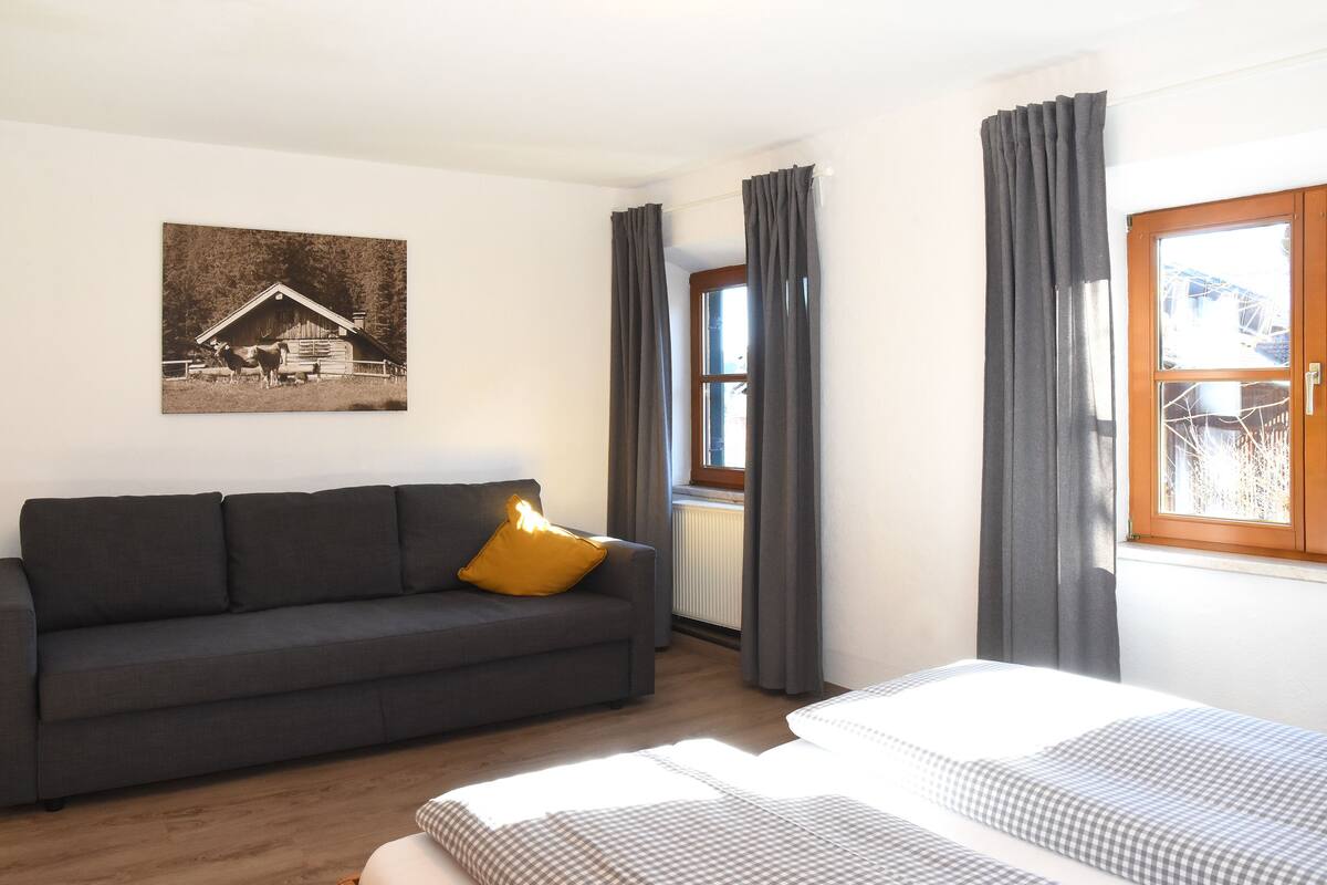 A room with a dark grey sofa, a bed with checkered bedding, a window with grey curtains, and a wall-mounted picture of a barn.