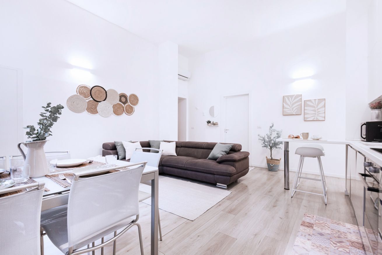 A modern, well-lit living room with a brown sofa, white dining table, and a kitchen area with a bar.