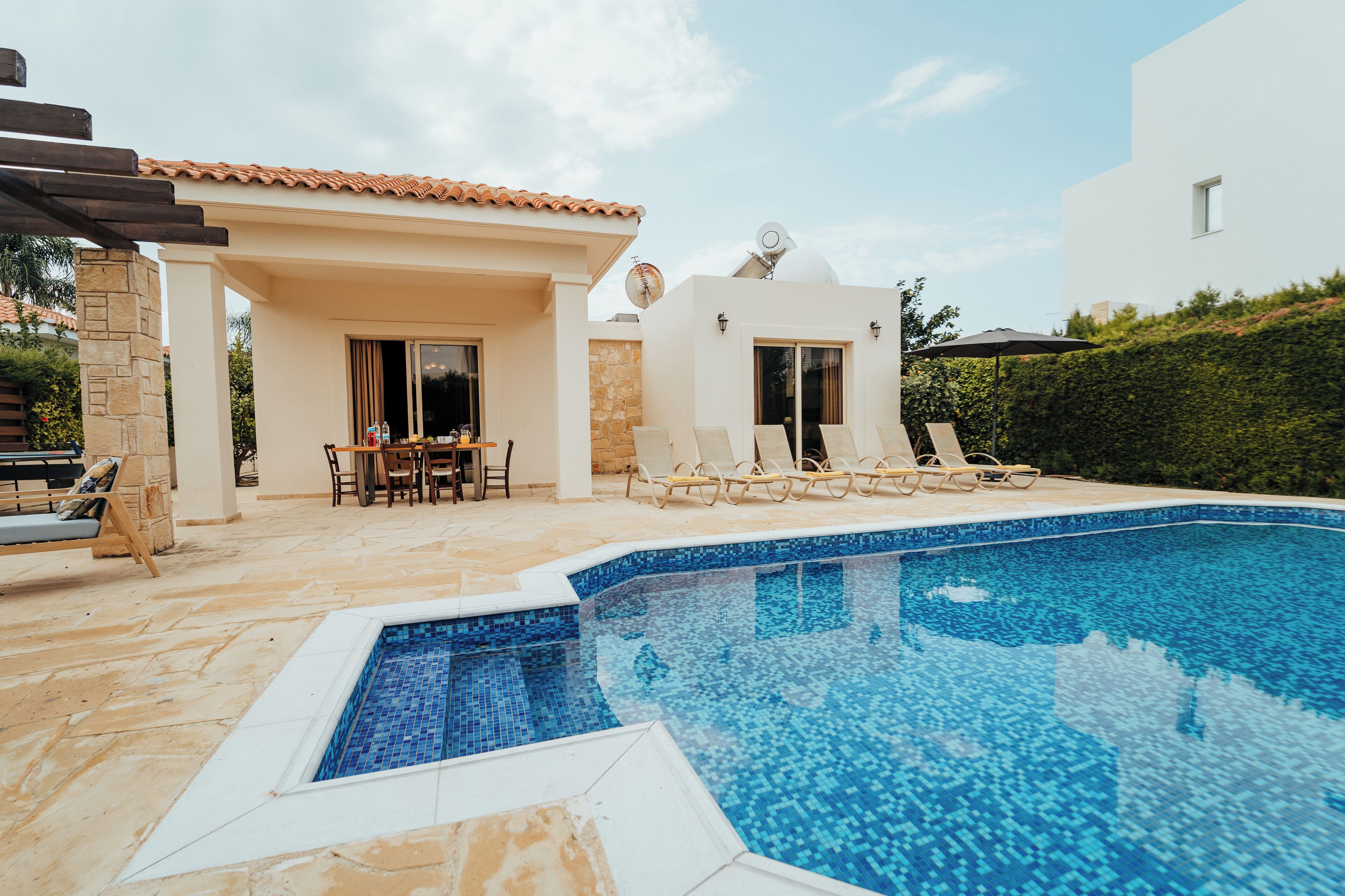 A swimming pool with lounge chairs, a covered patio area, and a white building with a tiled roof.