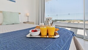 Une chambre d’hôtel avec un lit, un plateau de petit-déjeuner comprenant du jus d’orange, des fraises et des croissants, et une vue sur la plage.