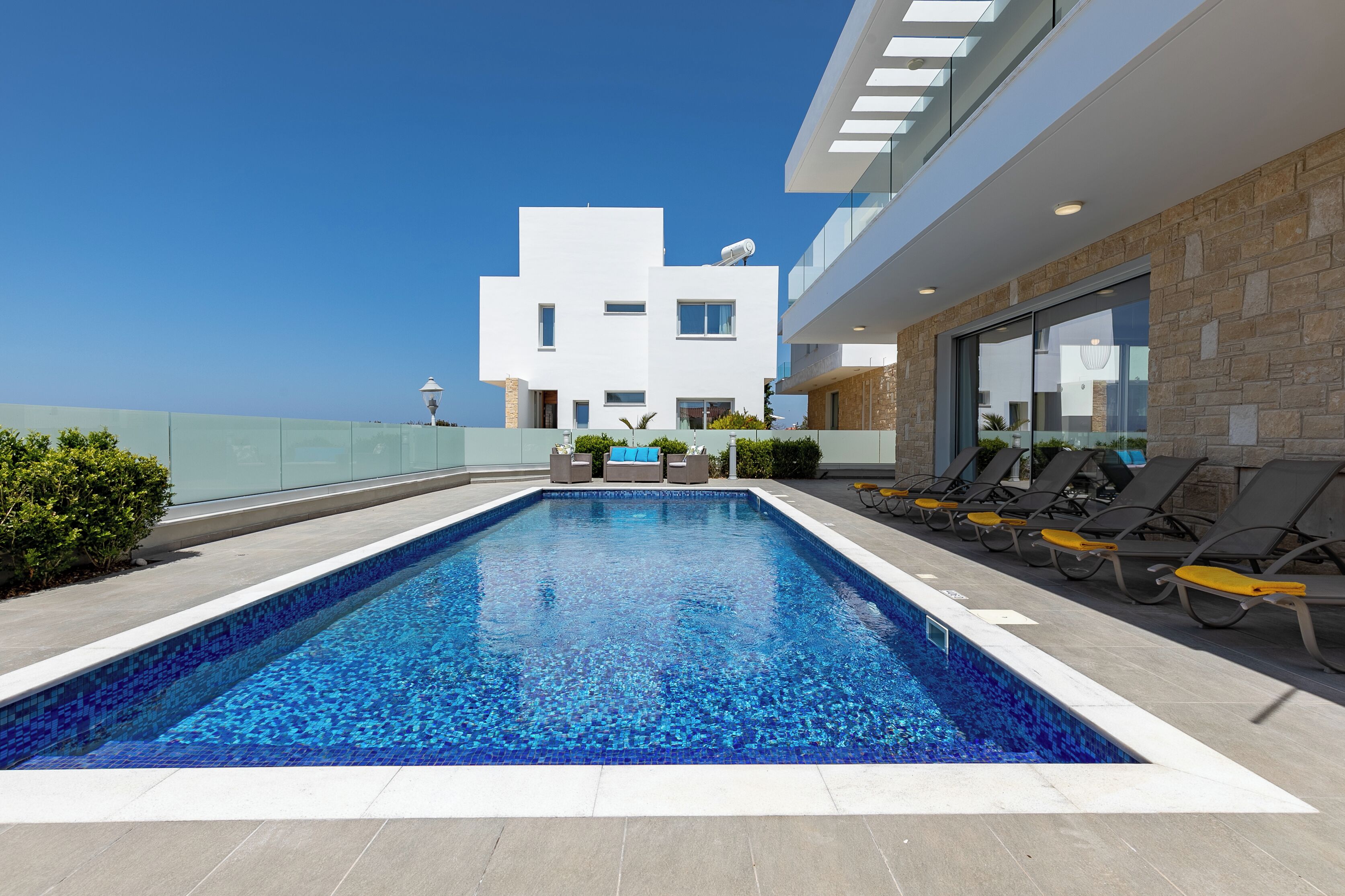 Un área de piscina moderna al aire libre con vista despejada al mar, un edificio blanco con techo plano y tumbonas a lo largo de la piscina.