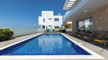 Un área de piscina moderna al aire libre con vista despejada al mar, un edificio blanco con techo plano y tumbonas a lo largo de la piscina.