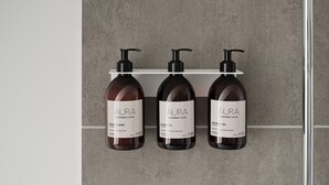 Tres botellas de gel de ducha AURA sobre un estante blanco frente a una pared con textura.
