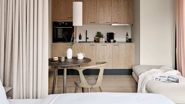 A modern hotel room with a dining area, kitchenette, and a bed with a magazine.