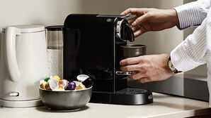 A person using a coffee machine in a kitchen with wooden cabinets and a stainless steel range hood.