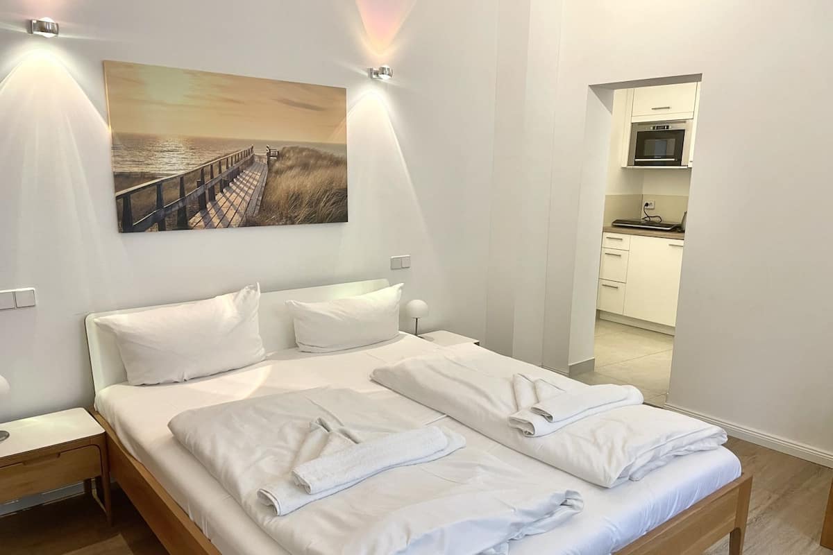 A neatly made bed with white linens and towels, a painting of a beach scene, and a view into a modern kitchen.