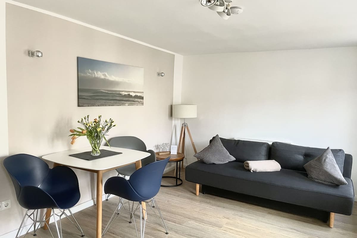 A modern living room with a dark grey sofa, two blue chairs, a round dining table with a vase of flowers, a floor lamp, and a painting of a seascape.