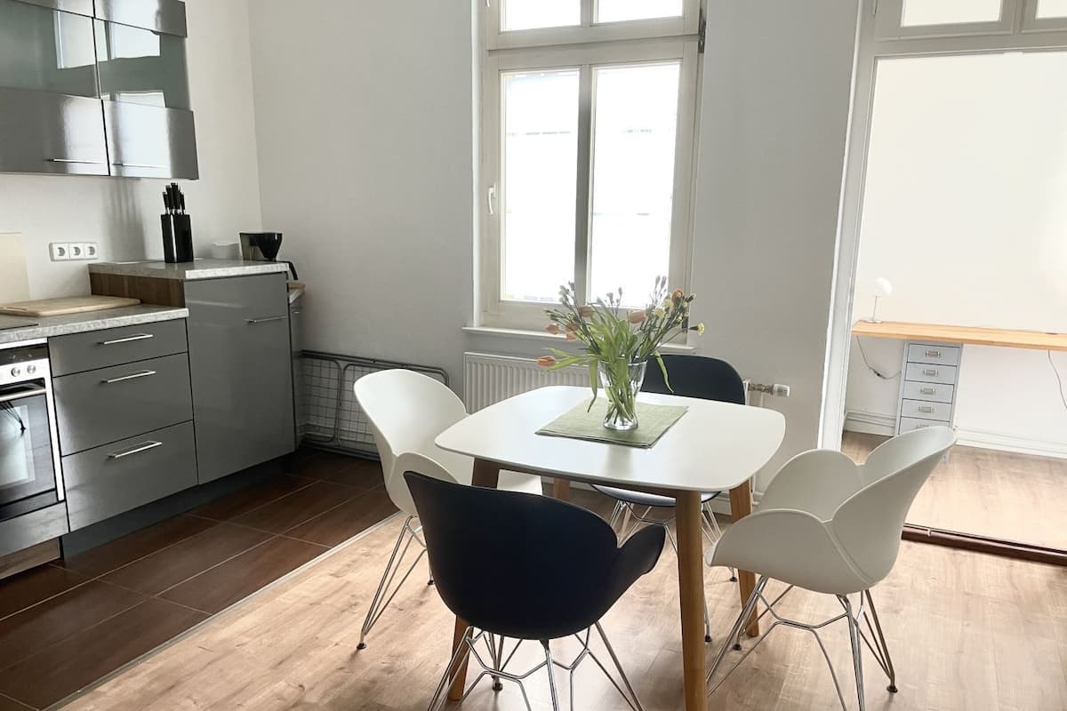 A modern kitchen with stainless steel appliances, a dining table with white chairs, and a window providing natural light.