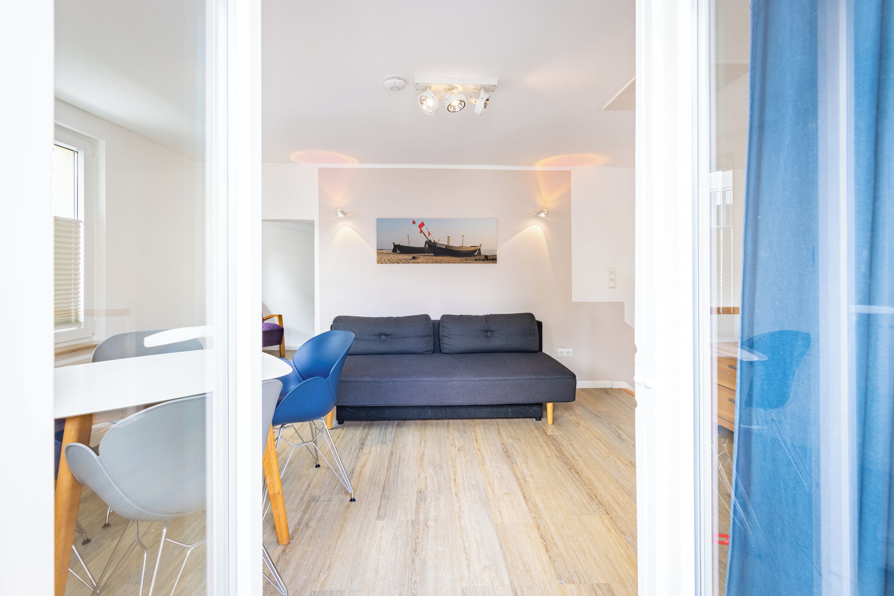 En moderne stue med sofa, spisebord og stole. Et billede hænger på væggen, og der er et vindue med persienner.