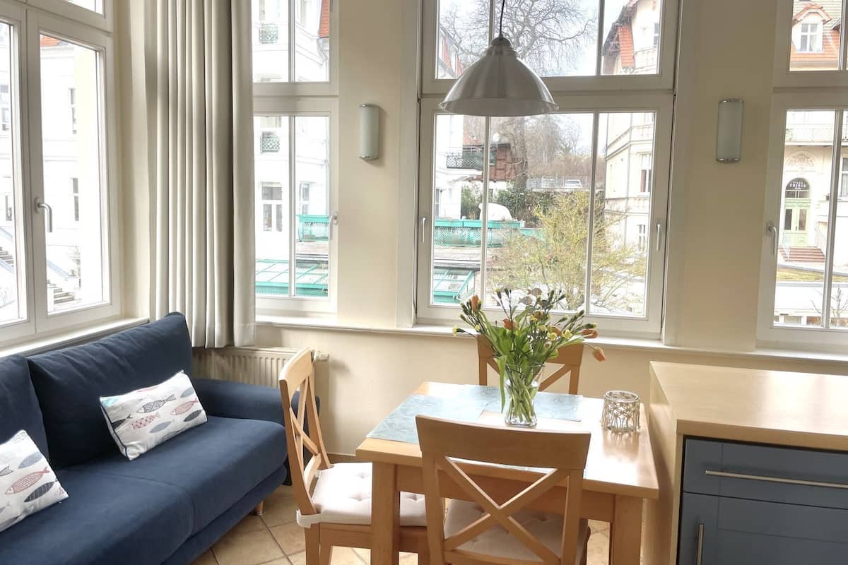 A cozy living room with a blue sofa, wooden dining table, and chairs. There is a vase with flowers on the table, and a hanging light fixture above.