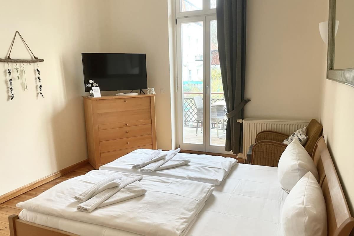A neatly made bed with white linens and towels, a wooden dresser with a television on top, and a window with a view of the outside.