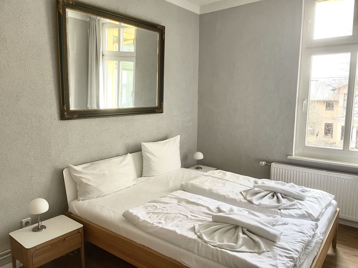 Une chambre à coucher comprenant un lit, une table de chevet, une lampe, un miroir et une fenêtre donnant sur des immeubles.